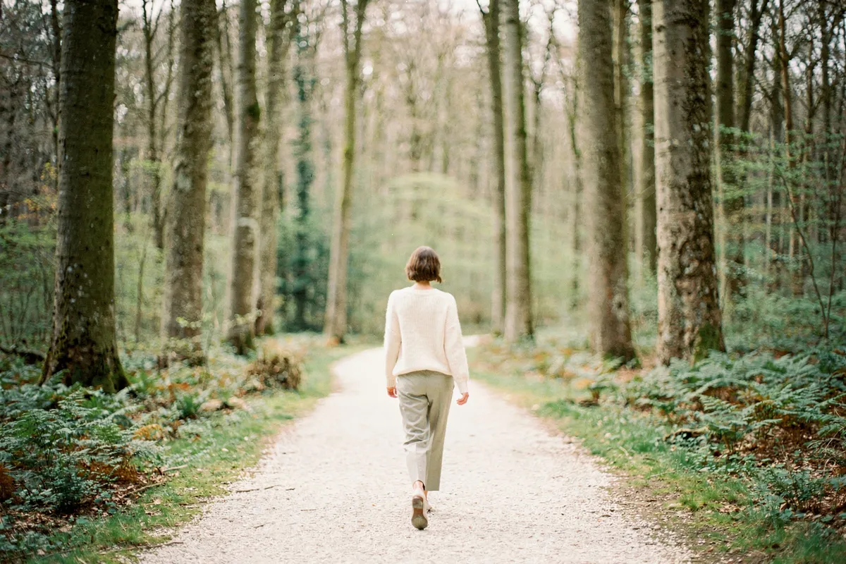 Une femme marchant lentement sur un chemin de gravier au milieu de grands arbres feuillus