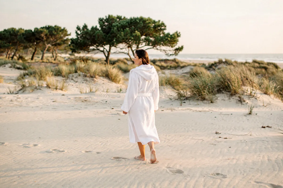 Une femme en peignoir blanc marche pieds nus sur une plage bordée de dunes et de pins